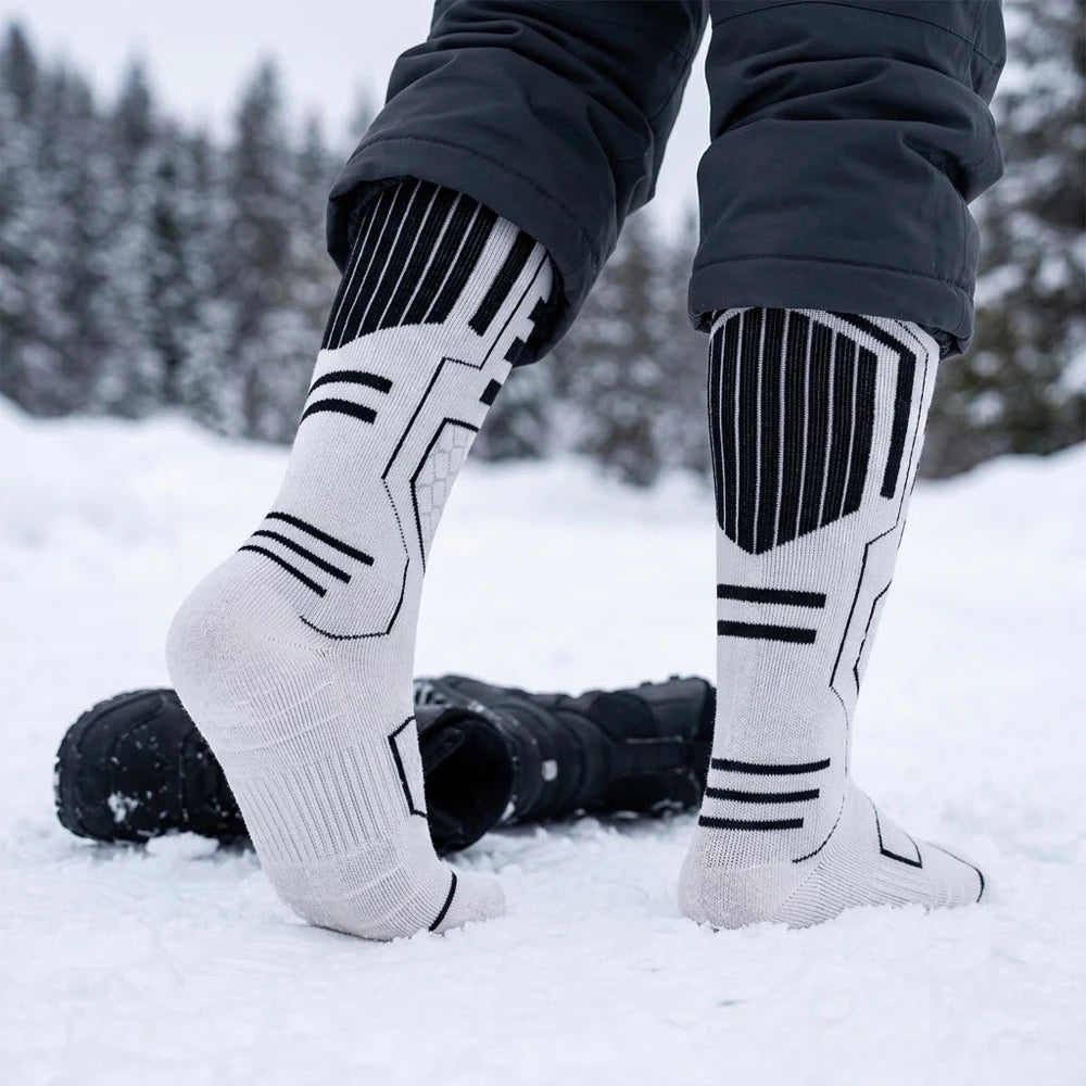 Feet wearing white Merino blend ski socks standing in the snow, with black winter boots resting nearby.