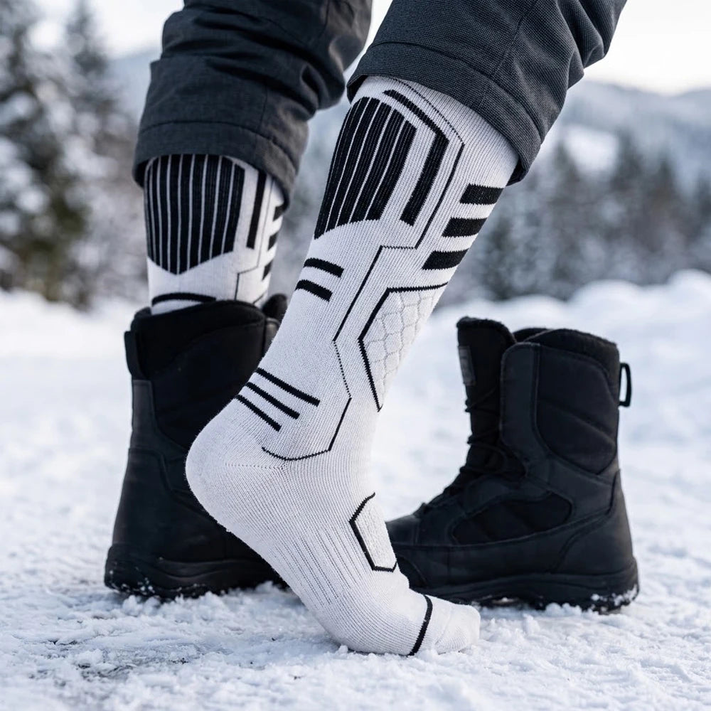 Person wearing white Merino blend ski socks on one foot and a black snow boot on the other, standing in the snow.